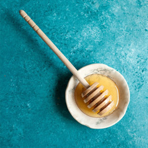 Auf einem türkisfarbenen Hintergrung ist eine kleine Schale mit Honig und einem typischen hölzernen Honiglöffel zu sehen. Das Bild steht für Hackerfalle Honeypot. Bild: www.unsplash.com / Photo by Lindsay Moe