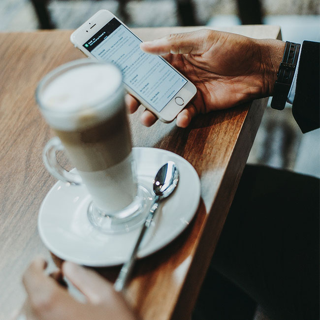 Zu sehen ist ein Tisch, auf dem ein Kaffee abgestellt ist, ein Geschäftmann sitzt davor und hält ein Handy in der Hand. Über mobile Anwendungen prüft er seine E-Mails. Bild: www.unsplash.com / Anete Lūsiņa