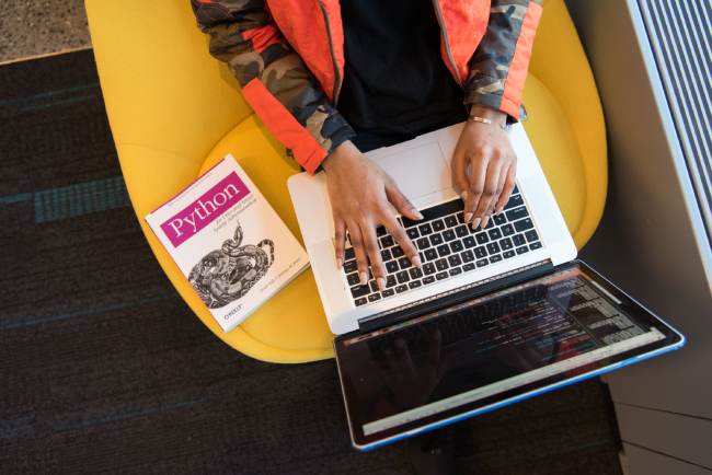 Hände bedienen einen Laptop, auf dem programmiert wird; ein Buch zur Programmiersprache Python liegt auf dem Tisch; wichtig ist, Security by Design mitzudenken. Bild: Pexels/Christina Morillo