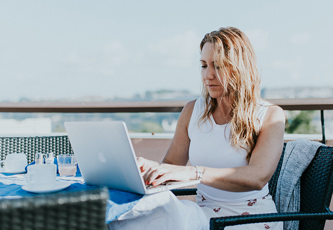 Eine Frau sitzt in einer Urlaubszene am Laptop und arbeitet. Sie nutzt das Workation-Konzept. Bild: Unsplash/Persnickety Prints
