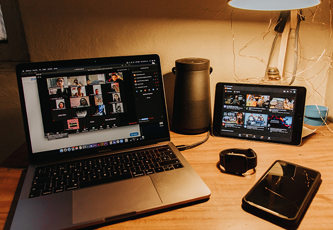 Zu sehen ist ein Schreibtisch mit Laptop und Tablet; darauf läuft ein Zoom-Meeting. Zoom bietet Live-Untertitel auf Englisch. Bild: Unsplash/Gabriel Benois