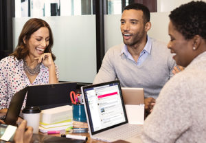 Ein Kollege und zwei Kolleginnen sitzen an einer Tischgruppe und unterhalten sich bei der Arbeit mit Office 2021. Bild: Microsoft