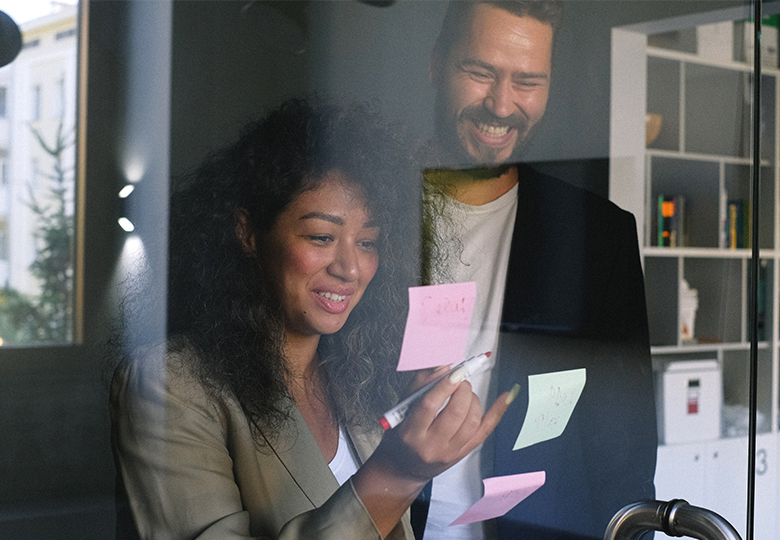 Eine Frau und ein Mann kleben Post-its an eine Glasscheibe; jetzt nutzen sie lieber Projekt-Management-Tools. Bild: Pexels/Anna Shvets