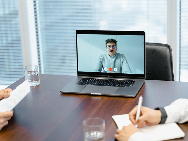 Zu sehen ist eine Videokonferenzszene aus dem Hybrid Office: Zwei Kollegen sitzen im Konferenzraum, ein Mann ist auf dem Laptop zugeschaltet. Bild: Pexels/MART PRODUCTION
