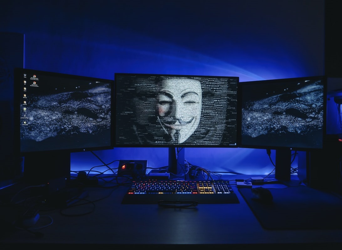 Drei Monitore, auf einem ist die Anonymous Maske der gleichnamigen Hackergruppe zu sehen. Bild: Unsplash/Stillness InMotion