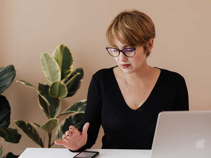 Frau am Laptop guckt auf ihr Smartphone. Thema des Artikels sind Phishing-Attacken via SMS