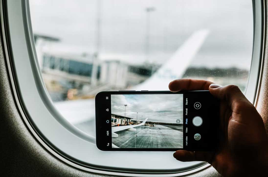 roaming-gebühren in der eu, es gilt seit Juli 2022 eine neue Verordnung. zu sehen ist die Hand einer Person, die mit dem Smartphone ein Foto im Flugzeug von den Tragflächen macht. Bild: Unsplash/Jonathan Kemper