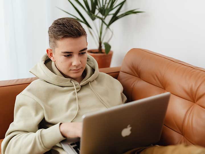 zu sehen ist ein Junge im Teenager-Alter, der mit seinem Laptop auf dem Sofa liegt. Das Thema sind Script-Kiddies, also jugendliche Amateur-Hacker. Bild: Pexels/Karolina Grabowska