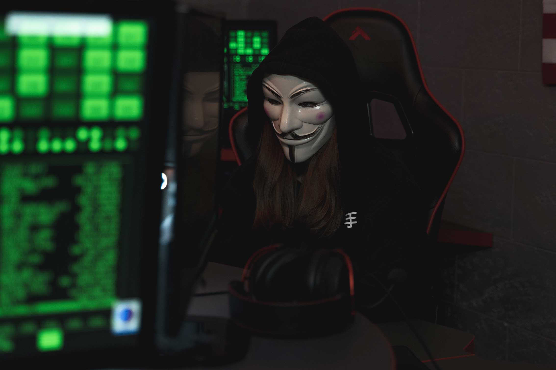 zu sehen ist ein klischeehafter Hacker mit Anonymous-Maske in einem dunklen Raum mit großen Monitoren. Das Thema ist Doxing. Bild: Pexels/Tima Miroshnichenko