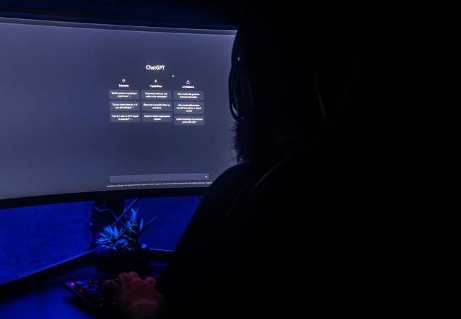 Zu sehen ist eine Person von hinten im Dunklen, die an einem PC ChatGPT nutzt - für KI-Angriffe? Bild: Pexels/Matheus Bertelli