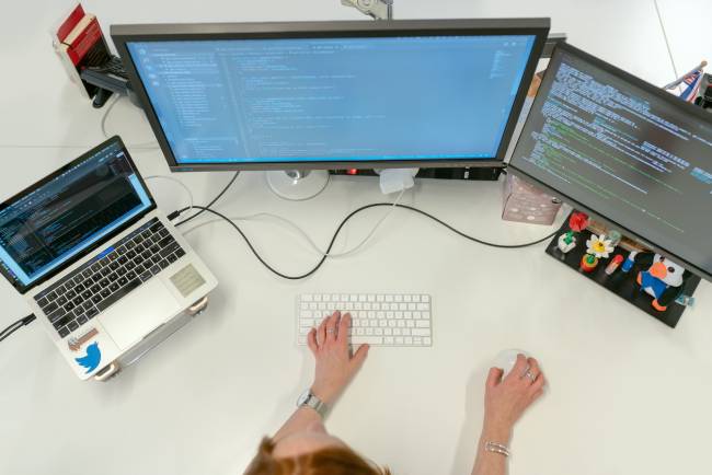 Zu sehen ist aus der Vogelperspektive ein Schreibtisch, an dem eine Frau an einem PV mit mehreren Bildschirmen an einer Software arbeitet; es geht um die Gefahr von Software-Supply-Chain-Attacken. Bild: Pexels/ThisIsEngineering