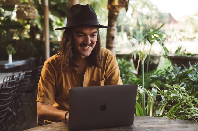 An Mann sitzt an einem tropischen Ort am Laptop und arbeitet; er nutzt das Modell Work from Anywhere. Bild: Unsplash/Matheus de Souza