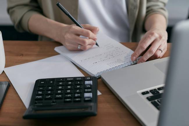 Zu sehen sind die Hände einer Frau, die mit Stift und Papier schreibt, ein Taschenrechner und Laptop. Es geht um die Pflicht zur E-Rechnung. Bild: Pexels/Mikhail Nilov