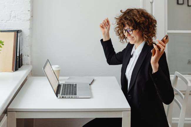 Zu sehen ist eine Frau am Laptop, die sich freut. Sie hat die Urlaubsübergabe fertig und macht jetzt den Laptop aus. Bild: Pexels/Karolina Kaboompics