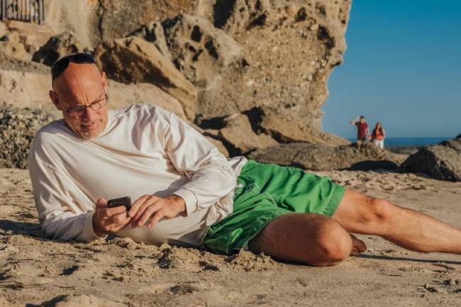 Ein Mann liegt mit dem Handy am Strand und wird von der Arbeit kontaktiert. Ein Fehler bei der Urlaubsübergabe? Bild: Pexels/Kindel Media