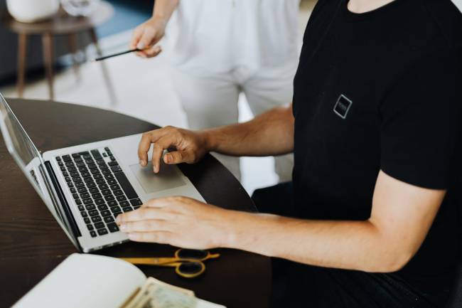 Zu sehen ist ein Mann am Laptop, eine Person steht daneben. Vielleicht sehen sie sich im neuen Outlook den Kalender an, um ein Meeting zu planen. Bild: Pexels/Karolina Kaboompics