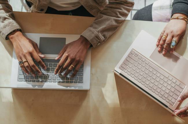 Zu sehen sind Hände und Laptops von zwei Personen auf einem Schreibtisch. Die Geräte könnten von der Outlook-Schwachstelle betroffen sein. Bild: Unsplash/Surface