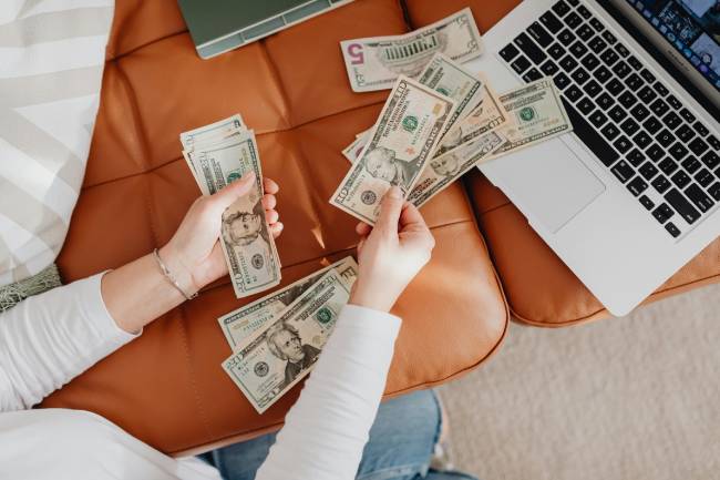 Zu sehen sind Hände die Dollar Scheine in der Hand haben und ein Laptop womöglich von Microsoft um die Kosten für die Updates vom Windows Server 2025 zu symbolisieren. Bild: Pexels/Kaboompics.com