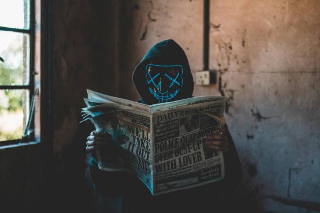 Zu sehen ist eine Person mit Maske auf und mit einer Zeitung in der Hand was einen Cyberkriminellen darstellen soll der Desinformation im Netz verbreitet. Bild: Pexels/Connor Danylenko