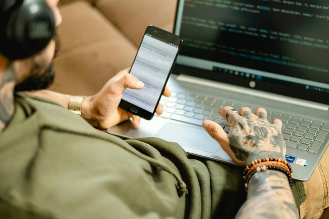 Zu sehen ist eine Person mit Handy vorm Laptop die womöglich gerade auf Fake Webseiten aufmerksam geworden ist. Bild: Pexels/Olha Ruskykh