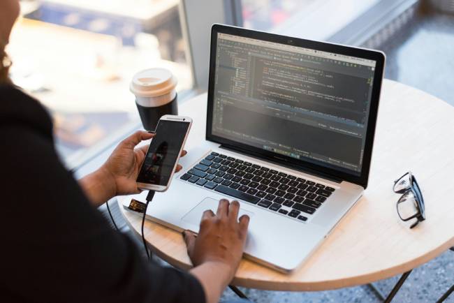 Zu sehen ist eine Frau vor einem Laptop und mit Handy in der Hand die wahrscheinlich Open Source Software nutzt. Bild: Pexels/Christina Morillo