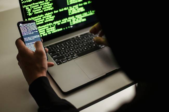 Zu sehen ist ein Hacker mit Laptop und Handy der womöglich gerade einen Hackerangriff durchführt und somit der Auslöser hybrider Bedrohungen ist. Bild: Pexels/Sora Shimazaki